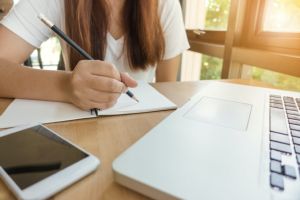Image of a student writing in a notepad next to a laptop.