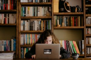 A young woman with curly hair studies at a laptop in front of a shelf of books. 
