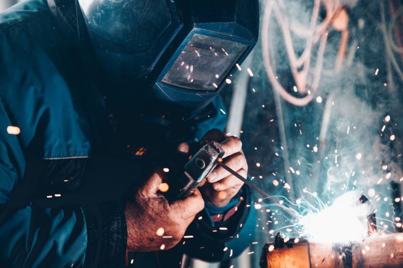 A welder in a face-covering safety mask works at welding with bright sparks. 