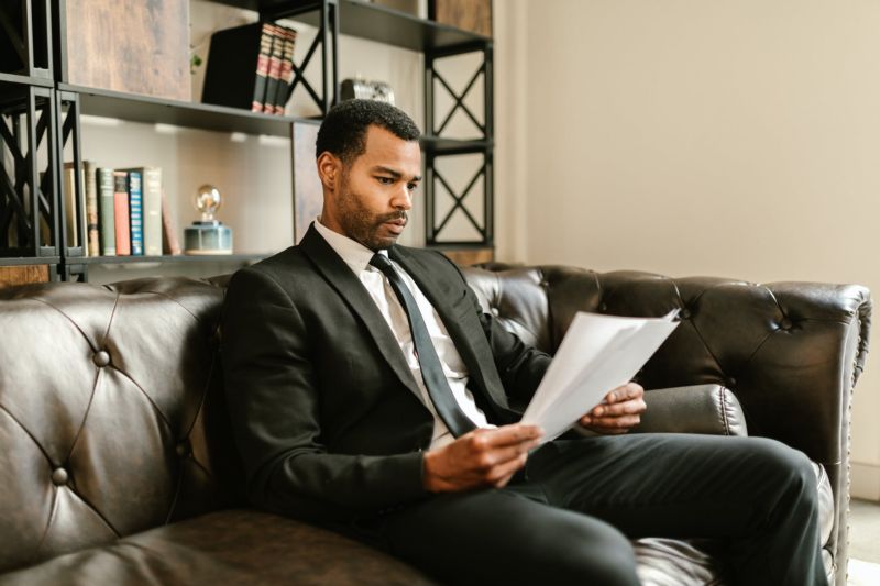 A man in a dark suit reads an article while sitting on a black leather couch. 