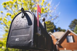 A black metallic mailbox  with red flag up, with a wooden house in the background. 